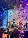 Band spelar på fargerik scene med konfetti over publikum i ein konsertsal.